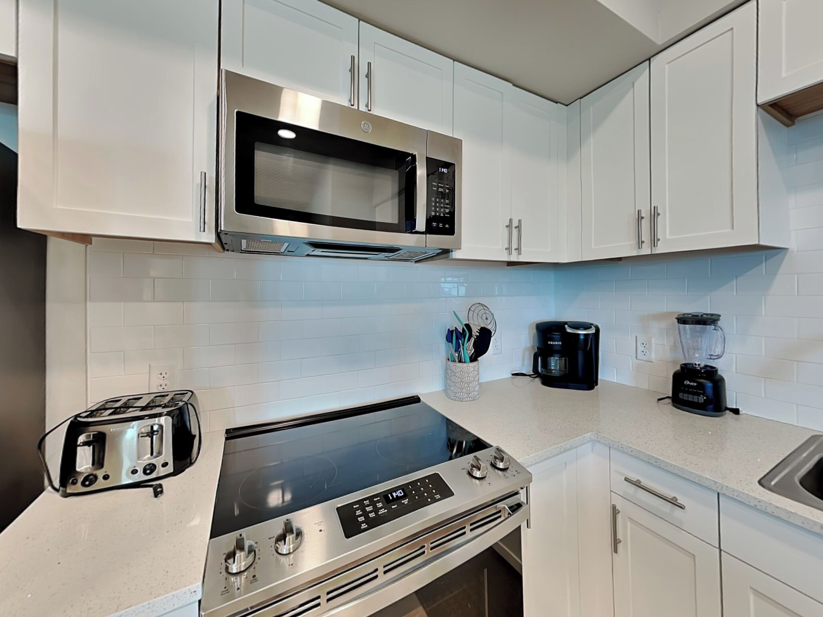 A modern kitchen with white cabinets, a stainless steel microwave, a toaster, a coffee maker, and a blender.