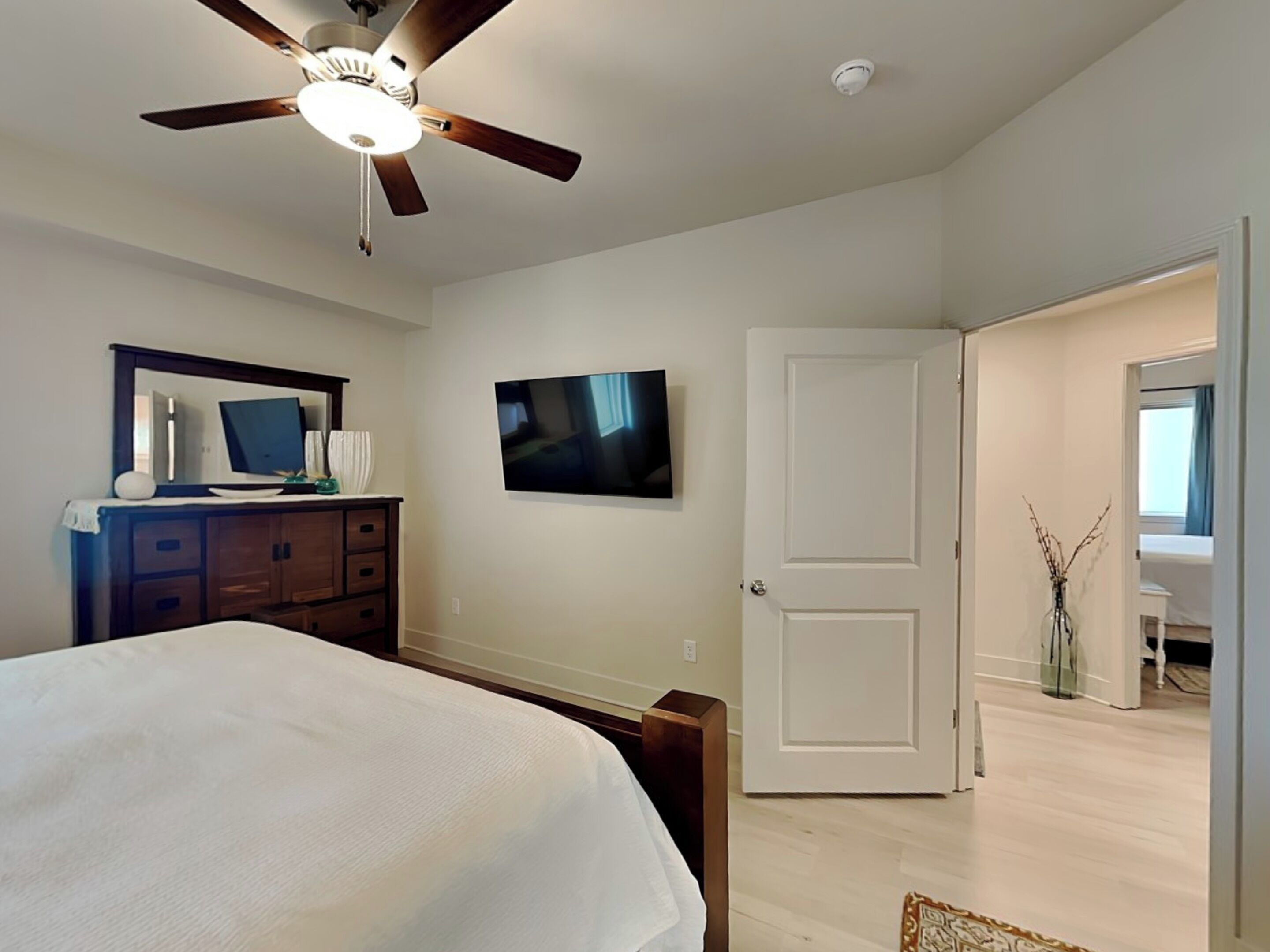 Un dormitorio con cama, cómoda, ventilador de techo, televisor y puerta que da a otra habitación.