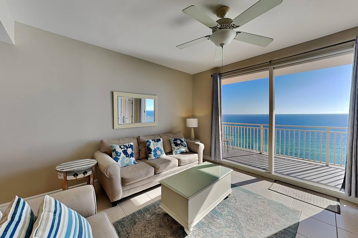 A living room with a beige sofa, striped pillows, a glass coffee table, a ceiling fan, and a large balcony with a view of the ocean.