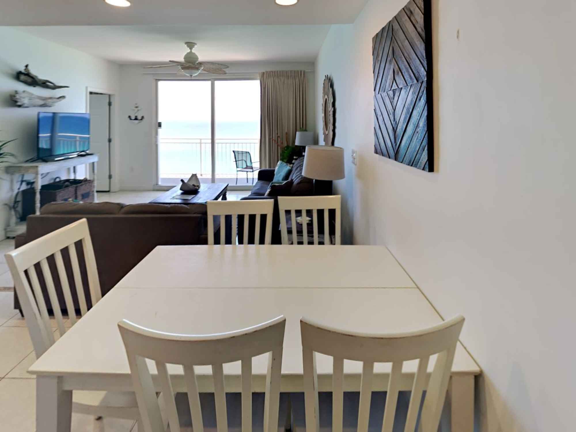 Un coin repas avec une table et des chaises blanches, un ventilateur de plafond et une vue sur l’océan par une grande fenêtre.