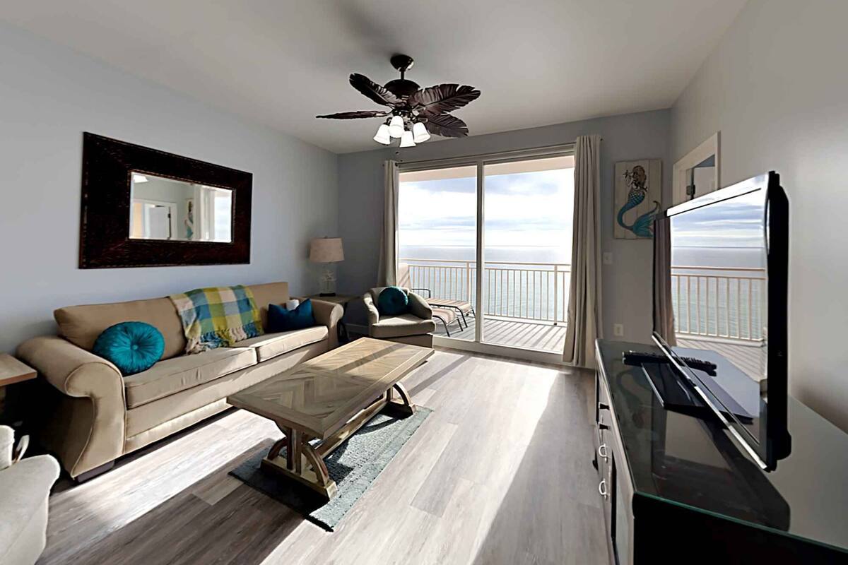 A living room with a sofa, coffee table, and a television. There is a balcony with a view of the ocean.