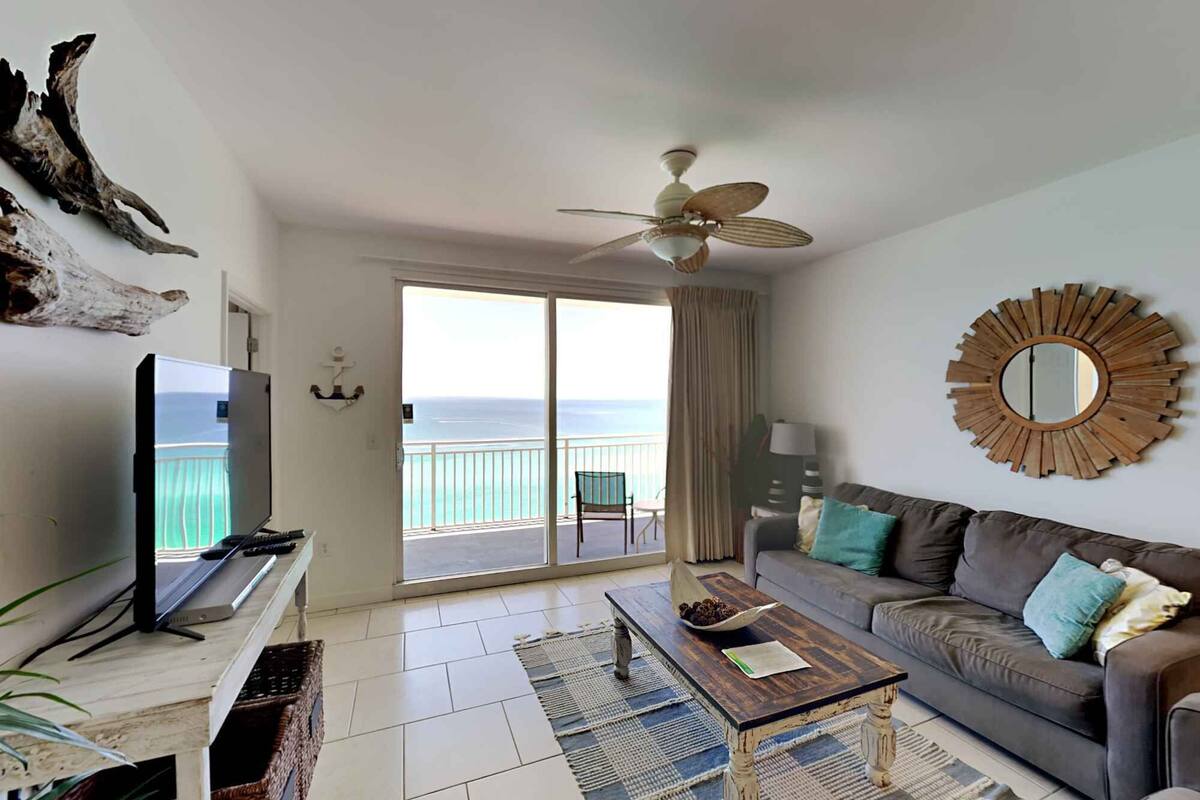 A living room with a TV, a ceiling fan, a sofa, a coffee table, and a view of the ocean through a sliding door.