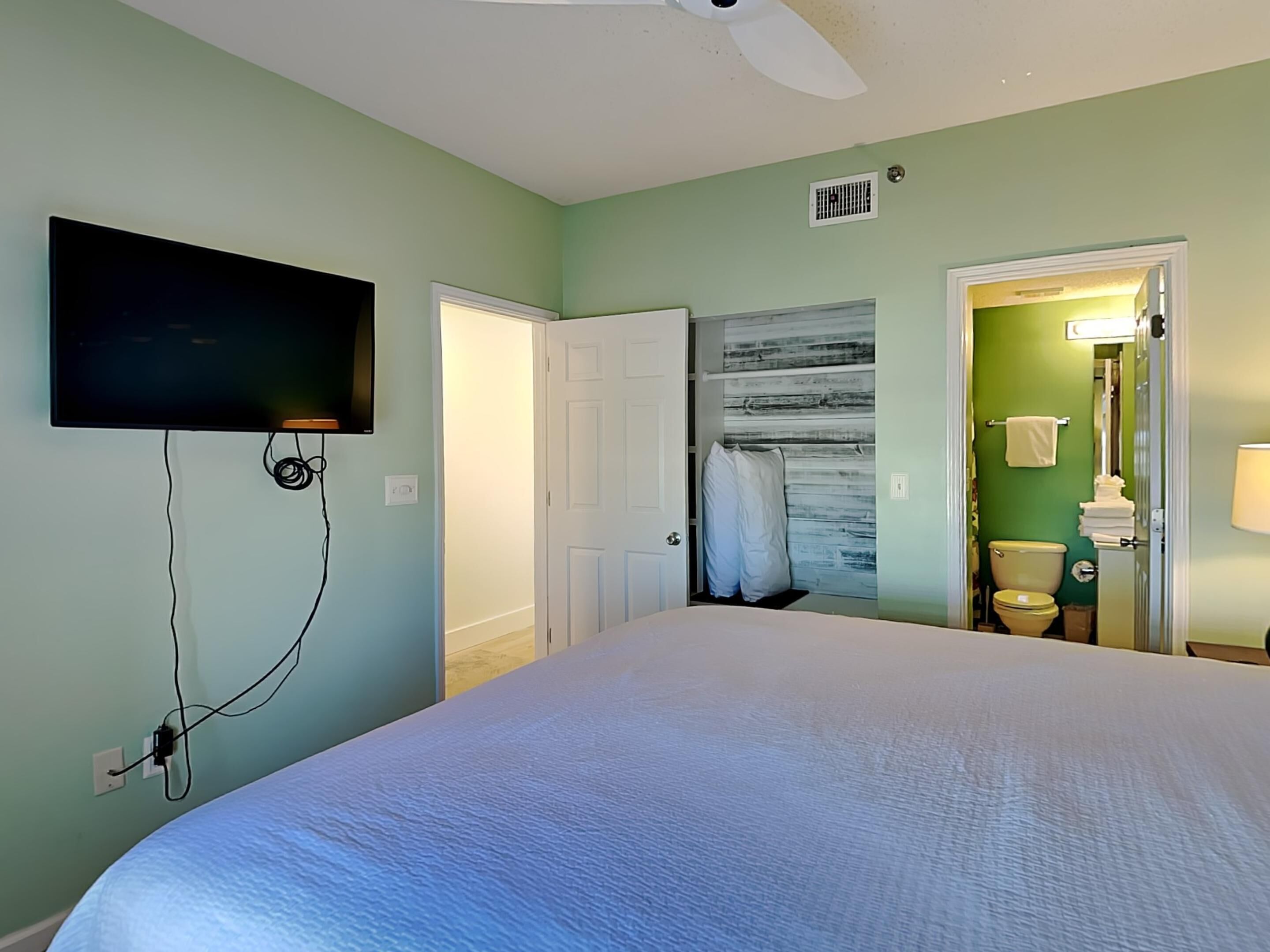 Une chambre à coucher avec un lit, un ventilateur de plafond, un téléviseur fixé au mur et une salle de bain visible à travers une porte ouverte.