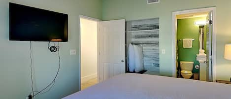 Un dormitorio con cama, ventilador de techo, televisor montado en la pared y baño visible a través de una puerta abierta.