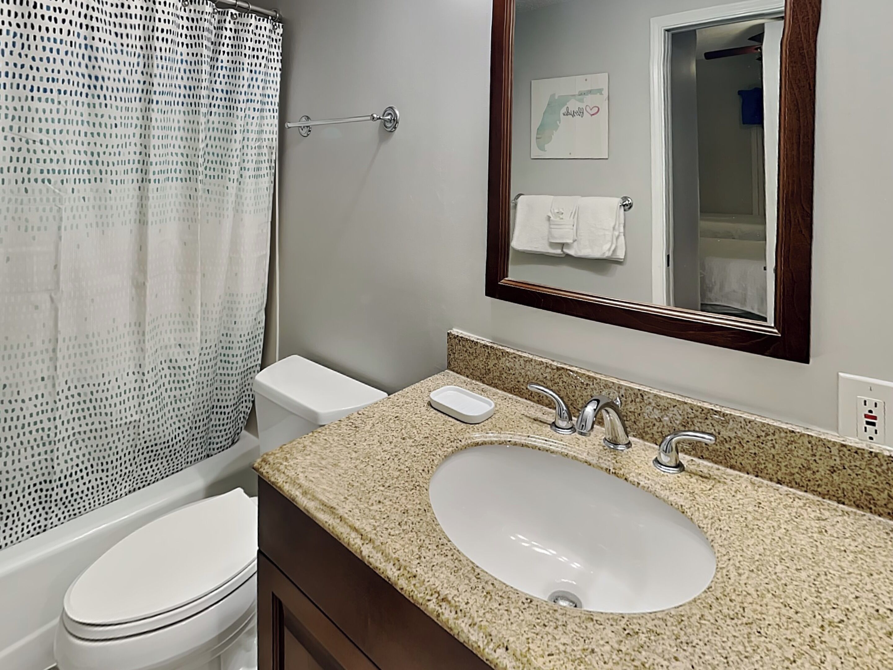 A bathroom with a white toilet, a sink with a granite countertop, a shower curtain, and a mirror with a towel hanging on it.