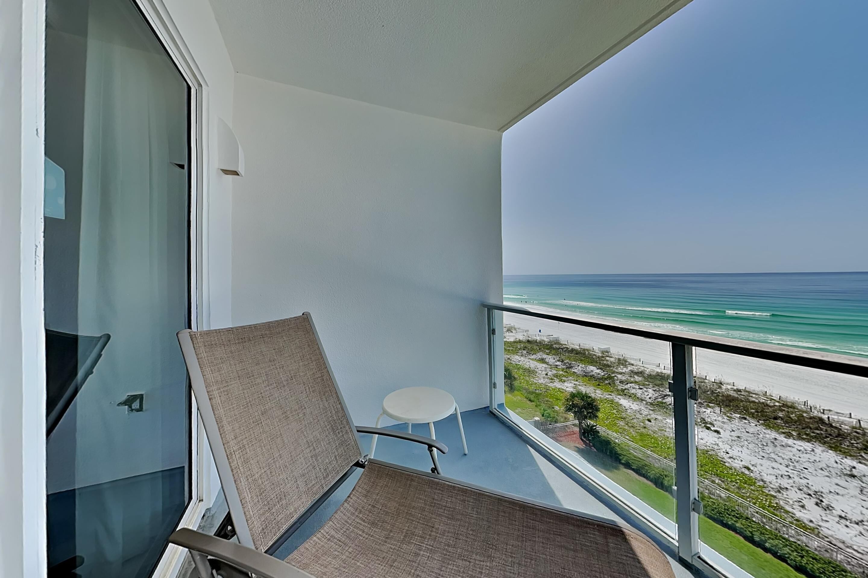 A balcony with a view of the beach, a white chair, and a small table.