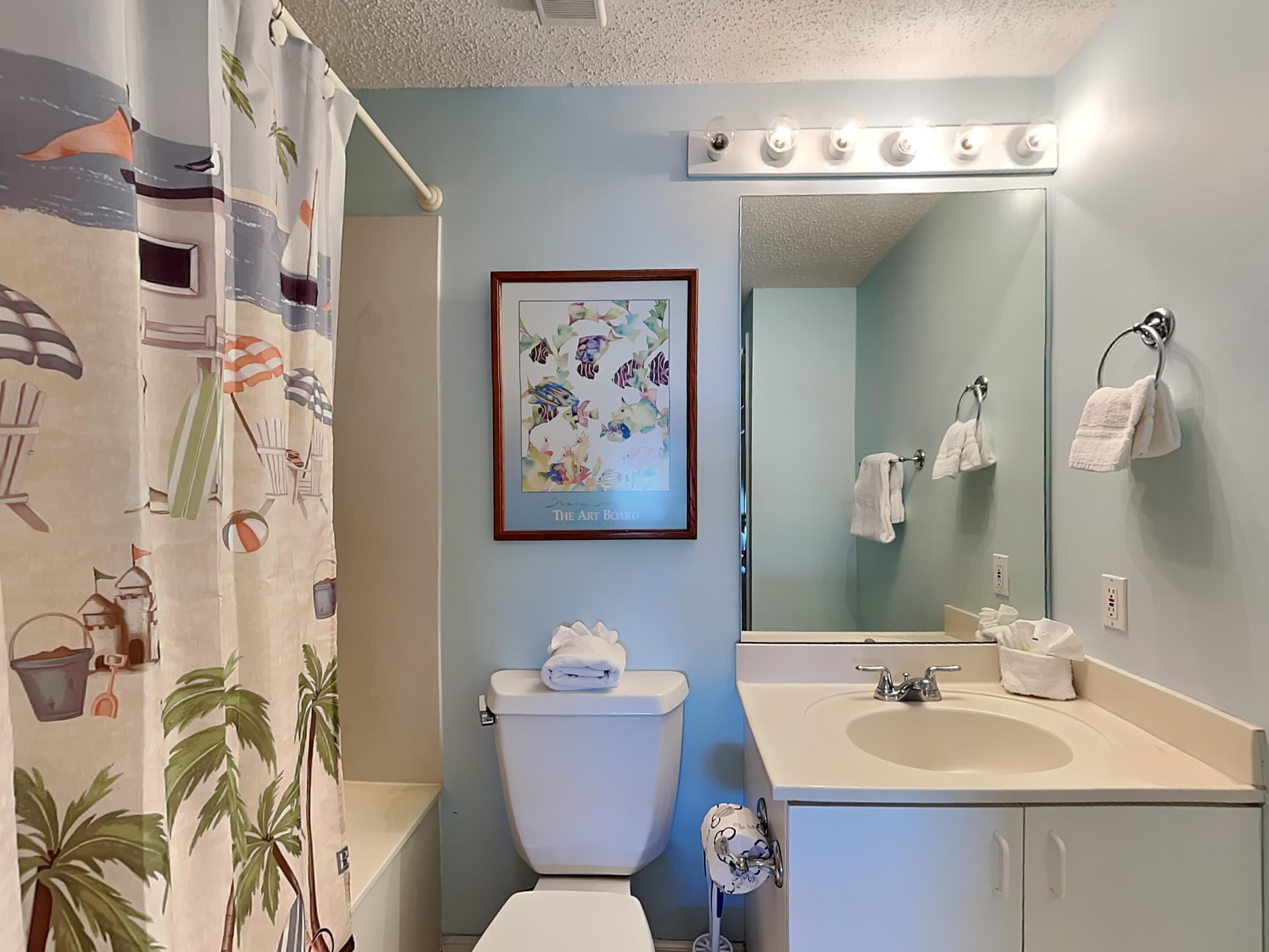 Une salle de bain équipée de toilettes, d’un lavabo et d’un miroir. On y trouve un rideau de douche à motifs de plage, une œuvre d’art encadrée accrochée au mur et un porte-serviettes avec des serviettes.
