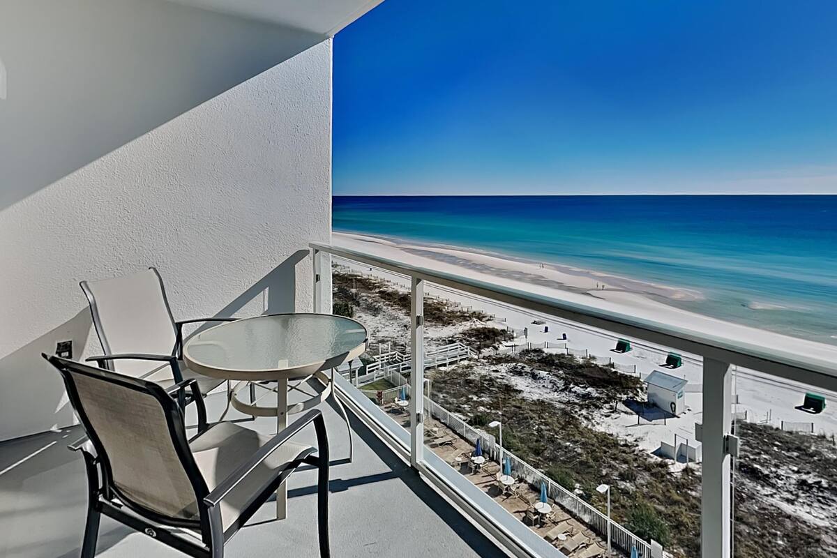 A balcony with a glass table and chairs, overlooking a beach with clear blue water and a sandy shore.