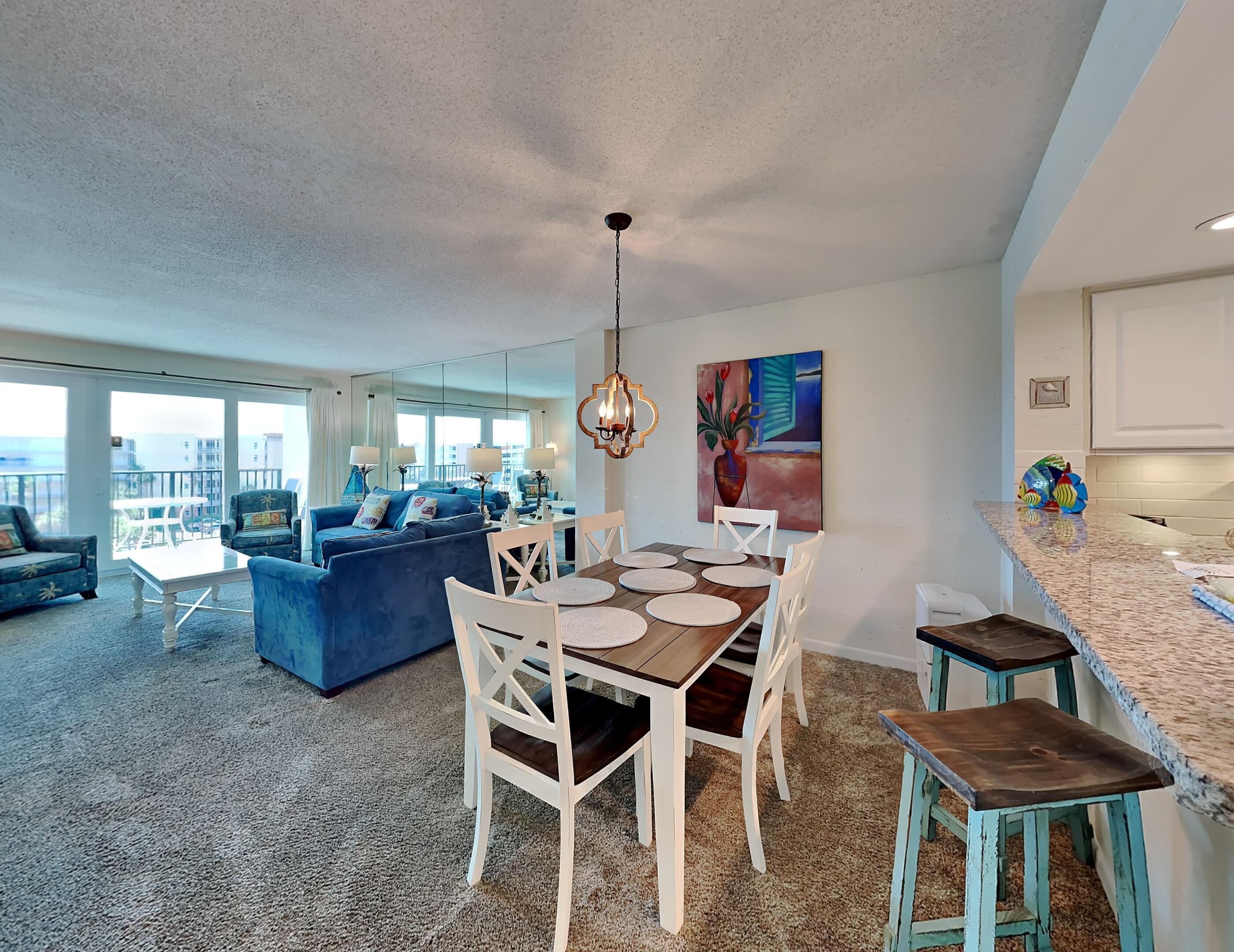 A modern living room with a blue sofa, a dining table with white chairs, and a kitchen area with a granite countertop.