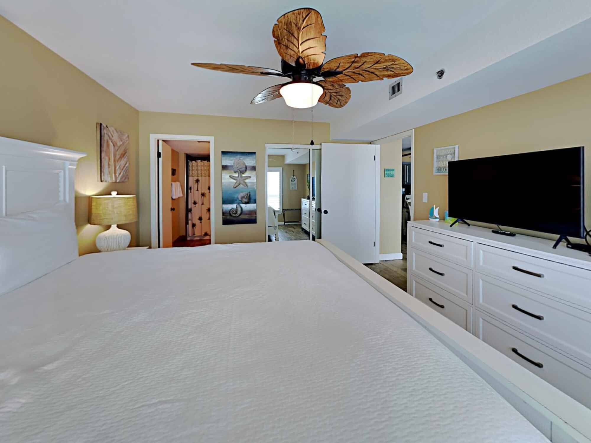 A bedroom with a large bed, a ceiling fan, a television, and a bathroom visible through an open door.