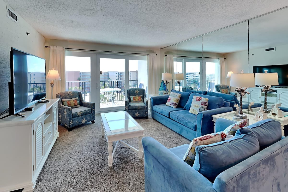 A modern living room with a large flat-screen TV, a blue sofa set, a coffee table, and a balcony with outdoor furniture.