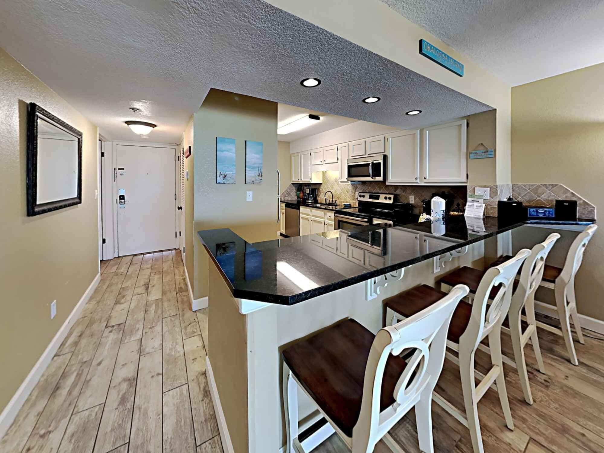 A modern kitchen with a central island, white bar stools, and a view of the hallway.