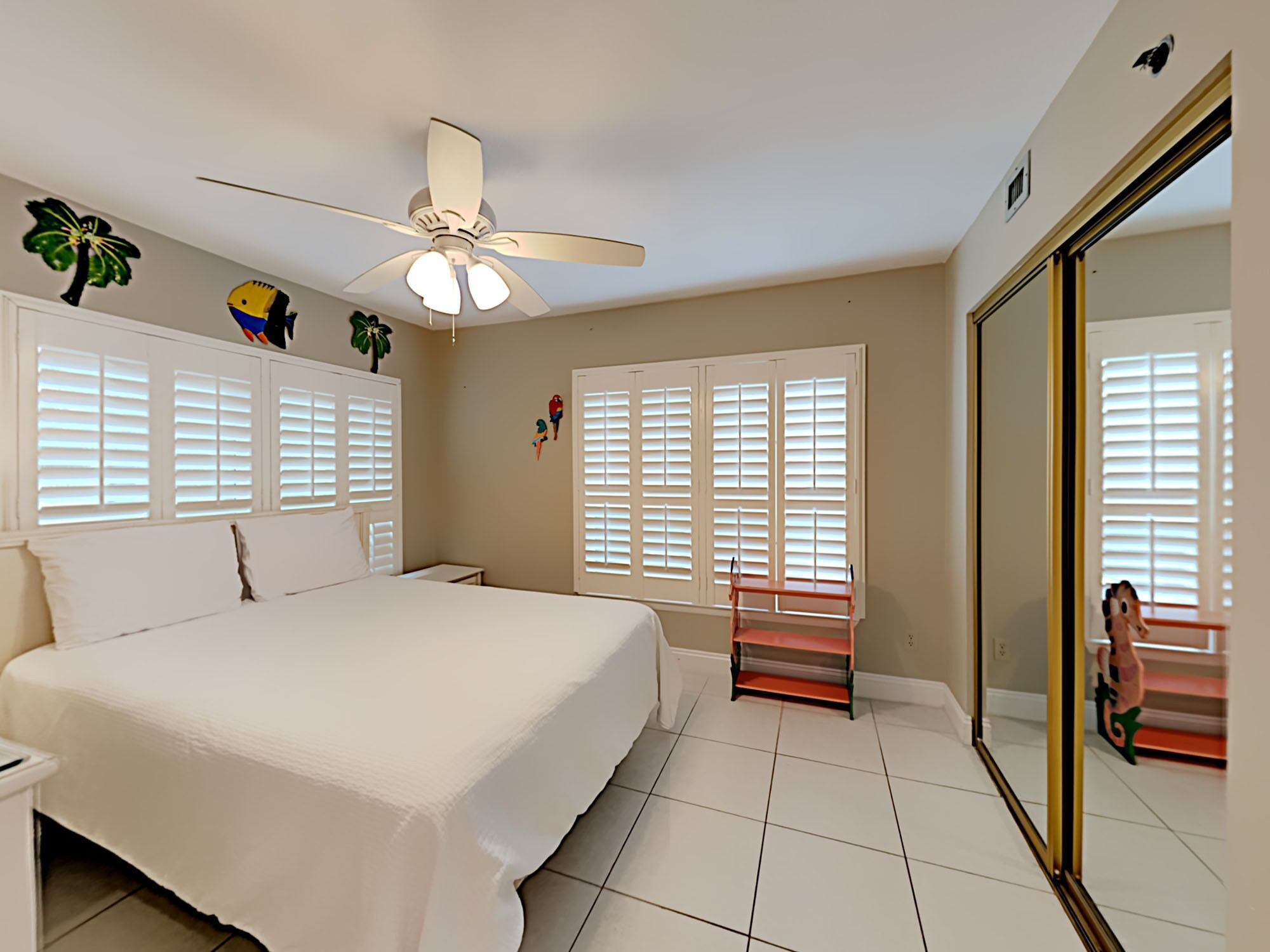 A bedroom with a bed, a ceiling fan, white shutters, and a large mirror.