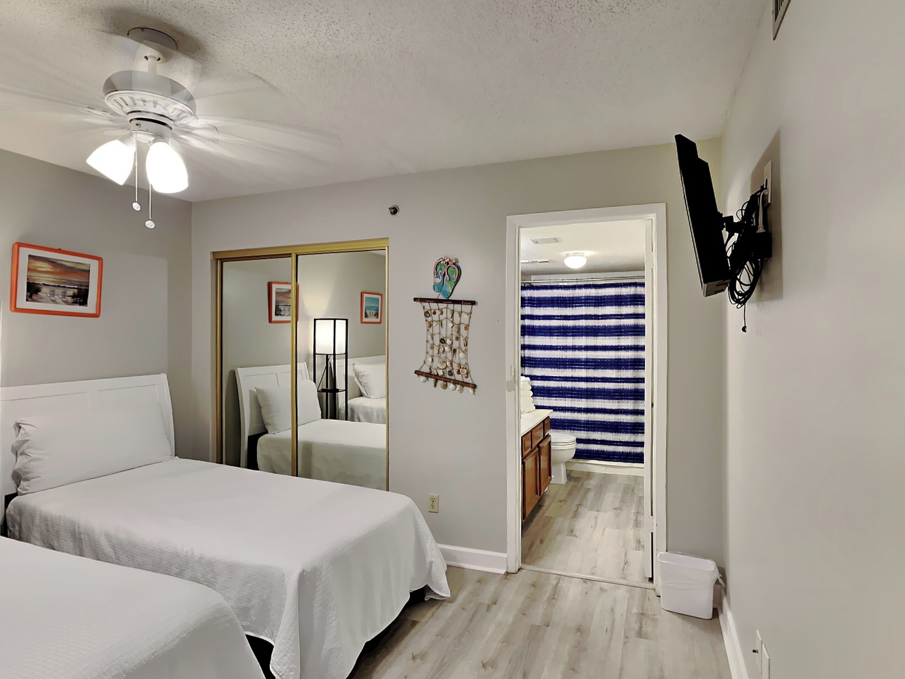 Ein Schlafzimmer mit zwei Betten, einem Deckenventilator, einem Spiegel und einem durch eine offene Tür einsehbaren Badezimmer.