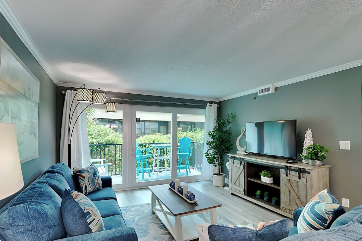 A modern living room with a sofa set, a coffee table, a TV on a wooden cabinet, and a balcony with outdoor furniture.