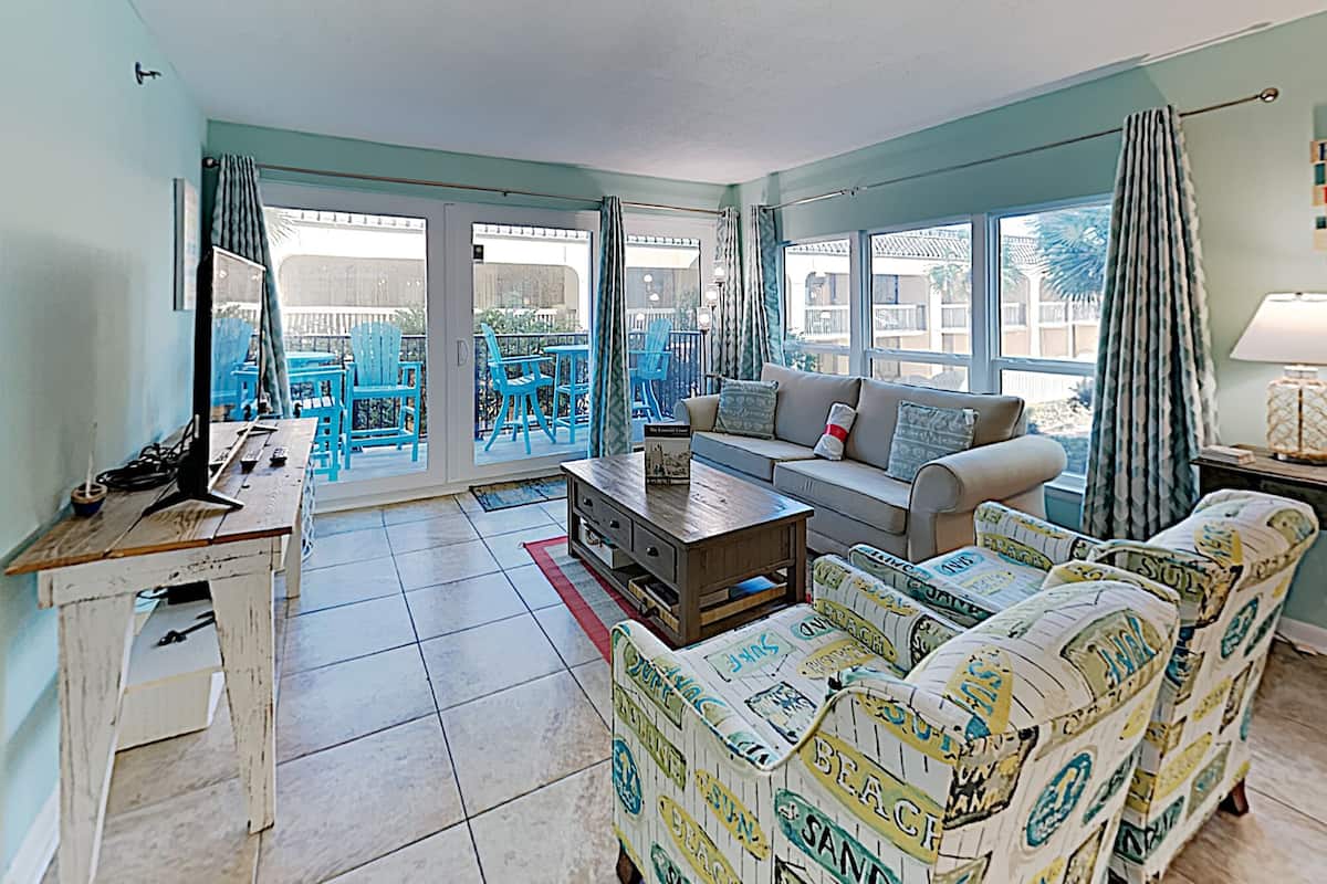 A living room with a sofa, coffee table, and a TV. There is a balcony with blue chairs and a table.