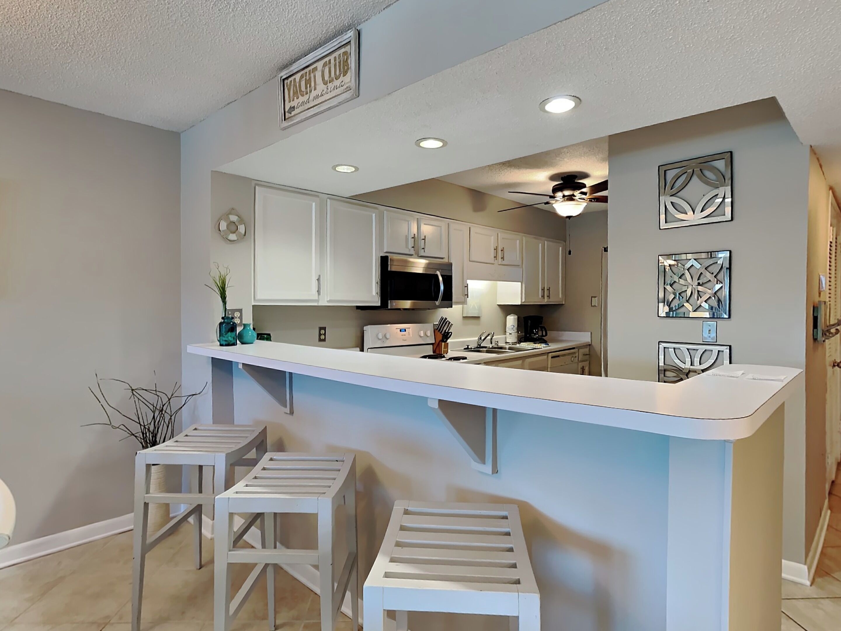 A modern kitchen with white cabinetry, a central island with stools, and a sign above reading "YACHT CLUB."