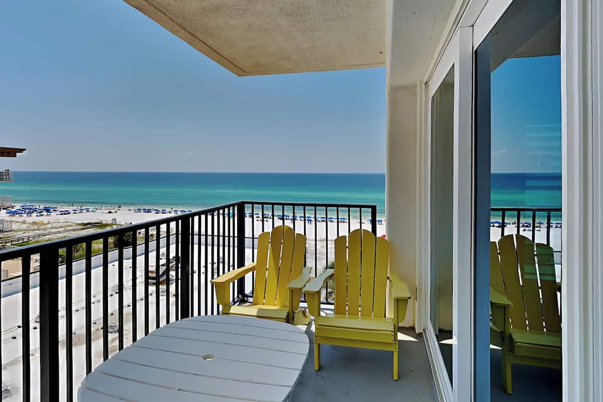 A balcony with two yellow Adirondack chairs and a white table, overlooking a beach with a clear view of the ocean and distant shoreline.