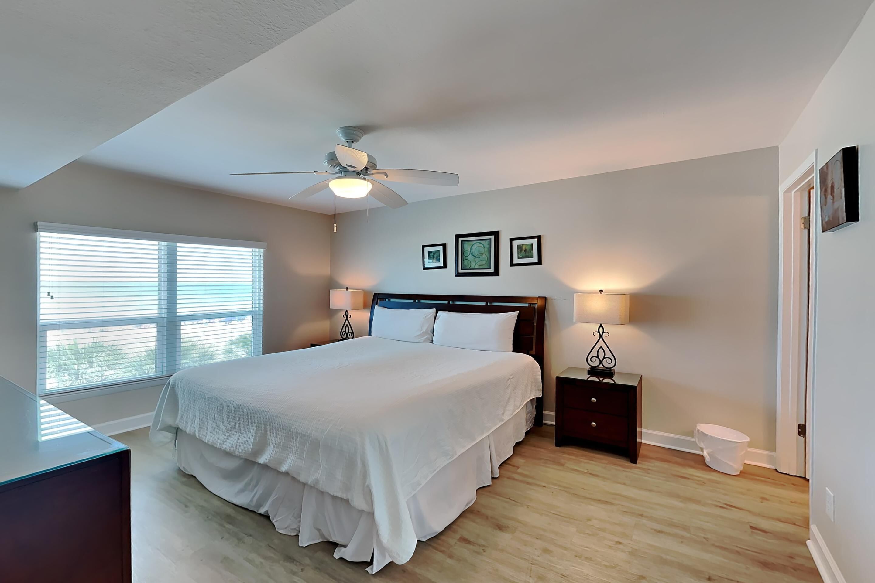 A bedroom with a large bed, a ceiling fan, a window with blinds, a bedside table with a lamp, and a small bedside cabinet.