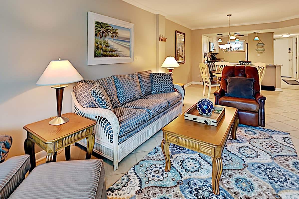 A living room with a patterned sofa, a coffee table, a red armchair, and a dining area in the background.