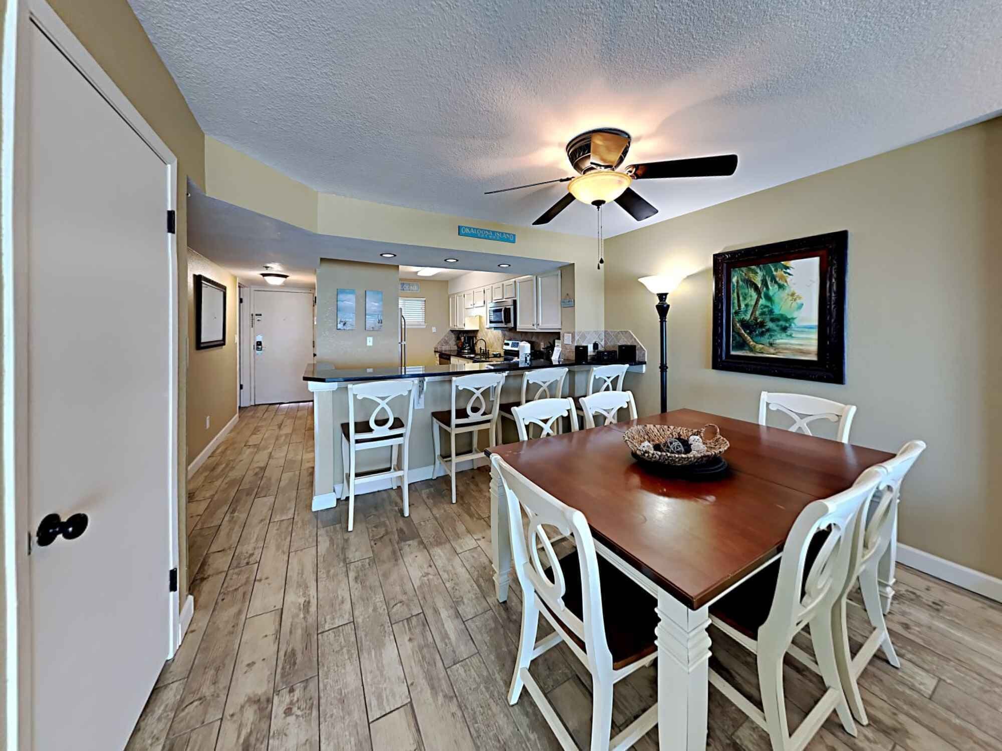 A dining area with a wooden table and white chairs, a kitchen in the background, and a ceiling fan.