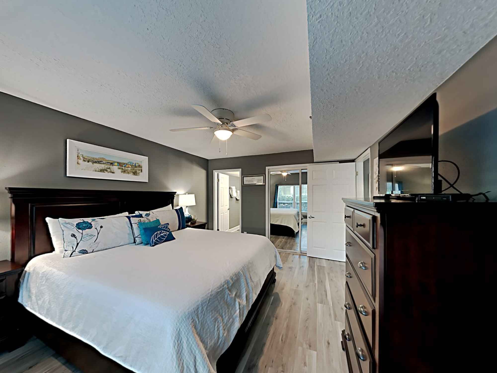 A bedroom with a bed, a nightstand, a dresser, a ceiling fan, and a door leading to another room.