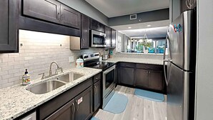 A modern kitchen with dark cabinets, stainless steel appliances, a granite countertop, and a view into the living area. - Sea Oats by Southern Vacation Rentals (Fort Walton Beach)