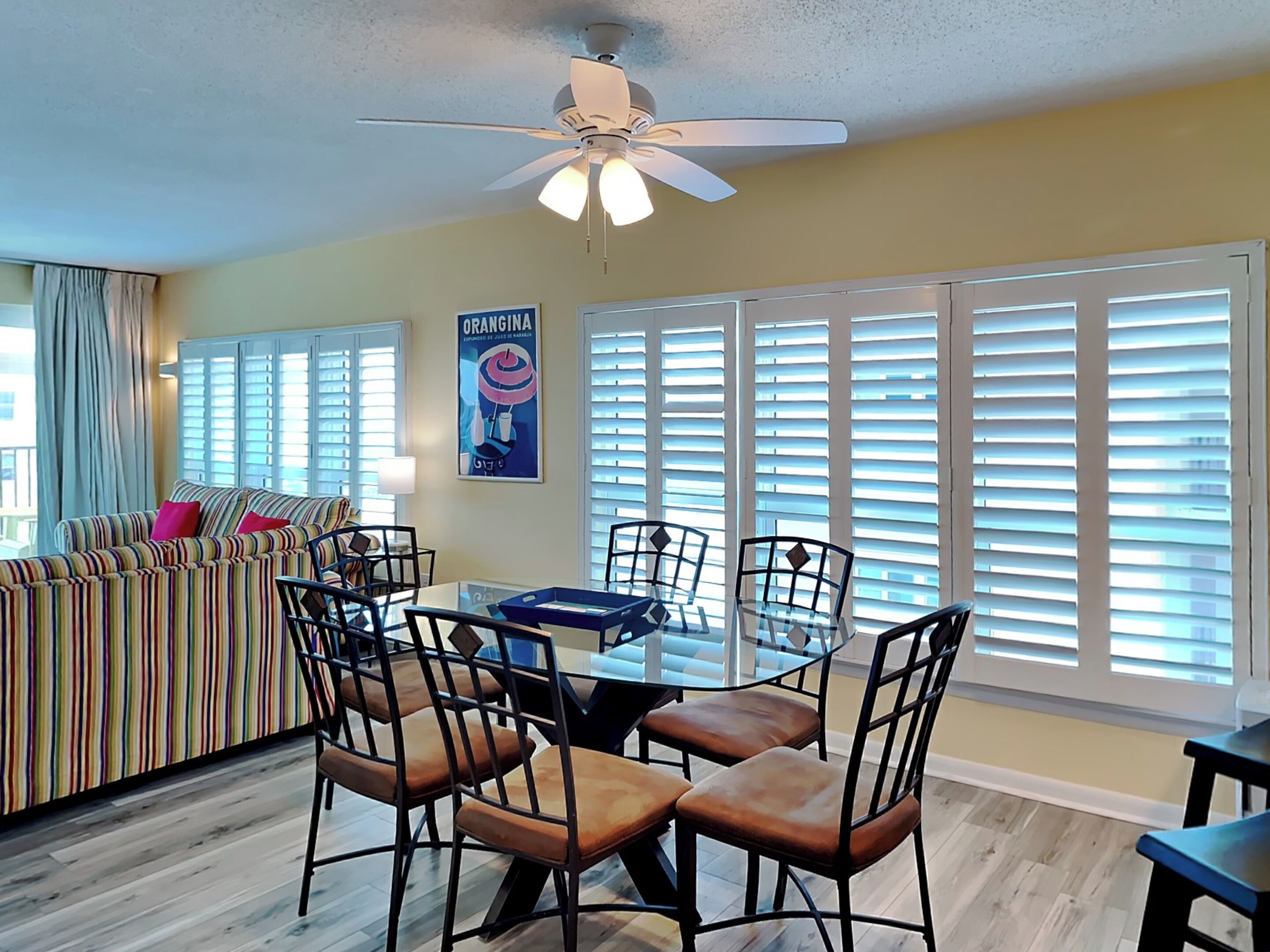 A dining area with a glass table and chairs, a ceiling fan, and large windows with blinds.
