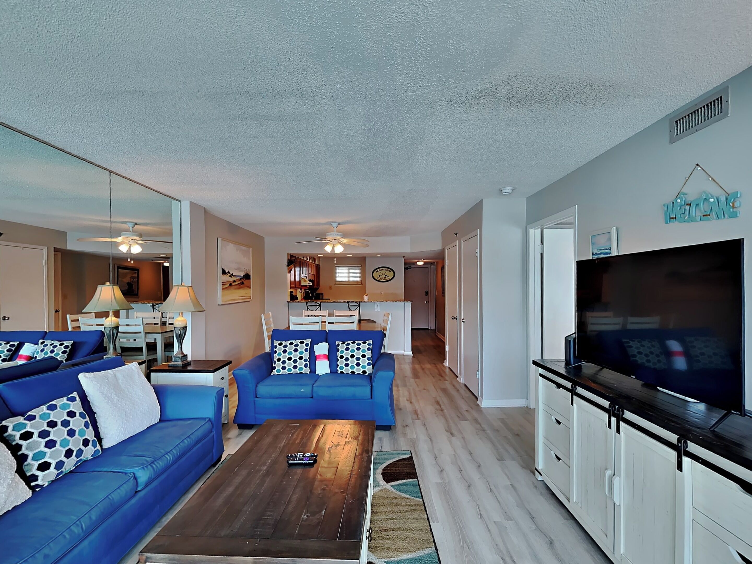A living room with two blue sofas, a coffee table, a TV, and a kitchen in the background.