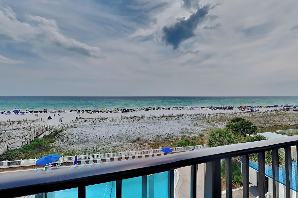 A beach view from a balcony with a pool, beach umbrellas, and lounge chairs.
