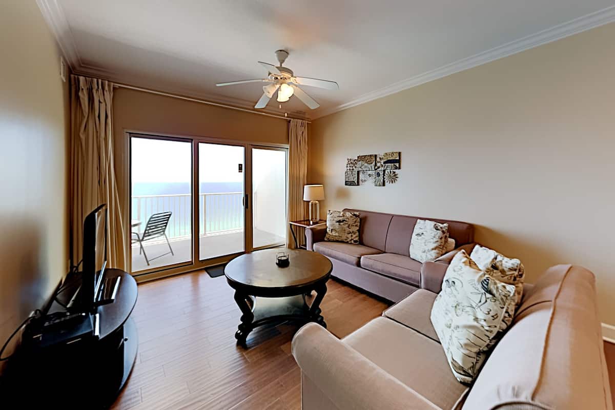 A living room with a sofa, a coffee table, a TV, and a ceiling fan. There is a sliding glass door leading to a balcony with a chair.