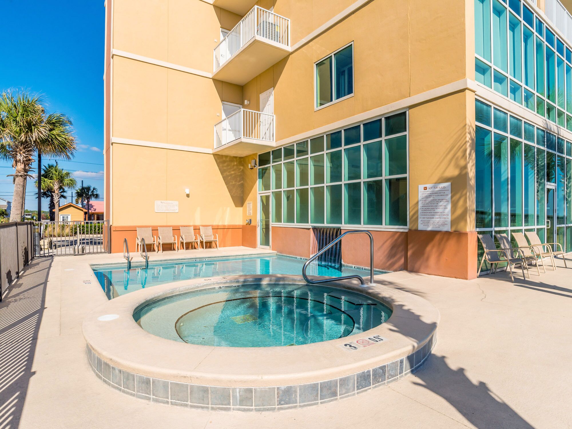A pool area with a hot tub, lounge chairs, and a building with large windows and a balcony.