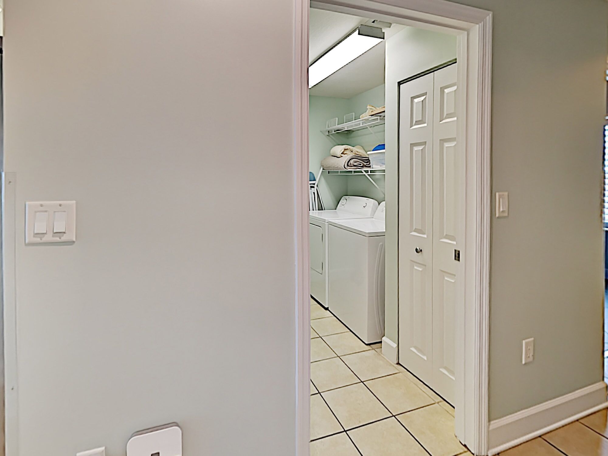A laundry room with a washing machine and dryer, a shelf with folded clothes, and a door leading to another room.