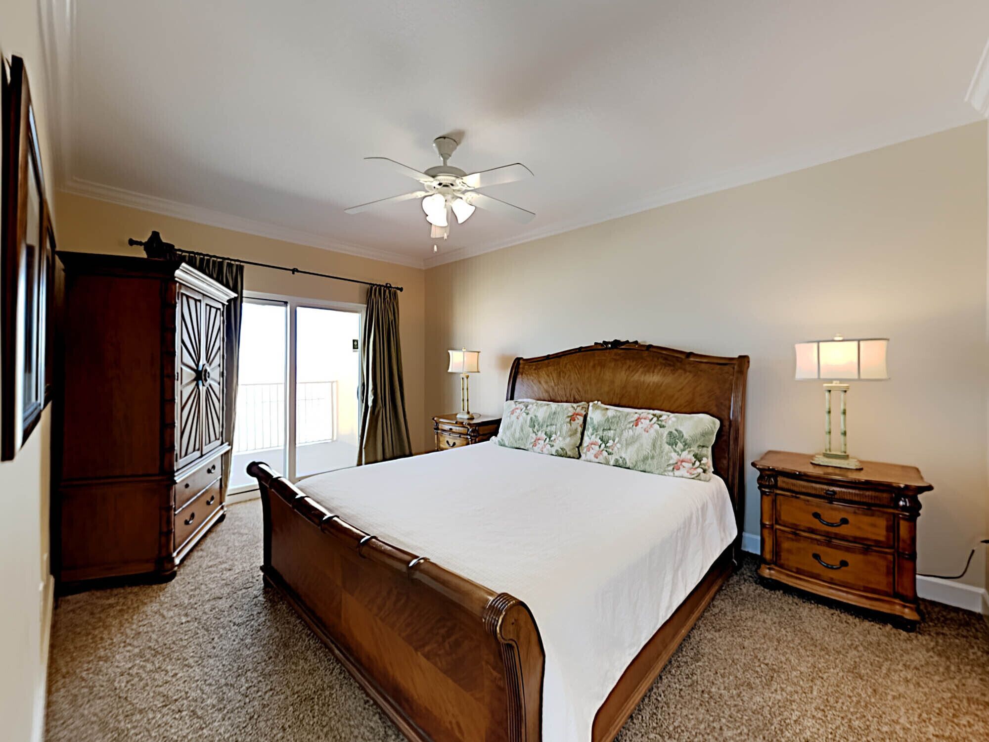 Quarto com cama grande, mesa de cabeceira de madeira com abajur, ventoinha no teto e vista para o exterior por uma porta deslizante.