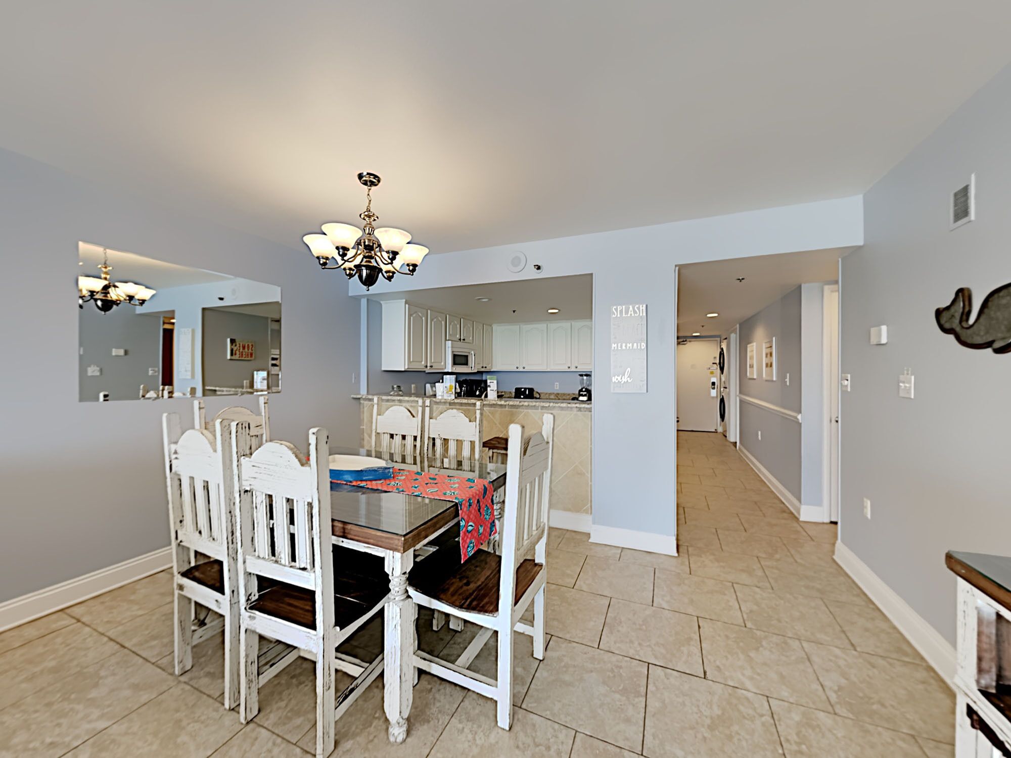 A dining area with a wooden table and white chairs, a kitchen in the background, and a hallway leading to another room.
