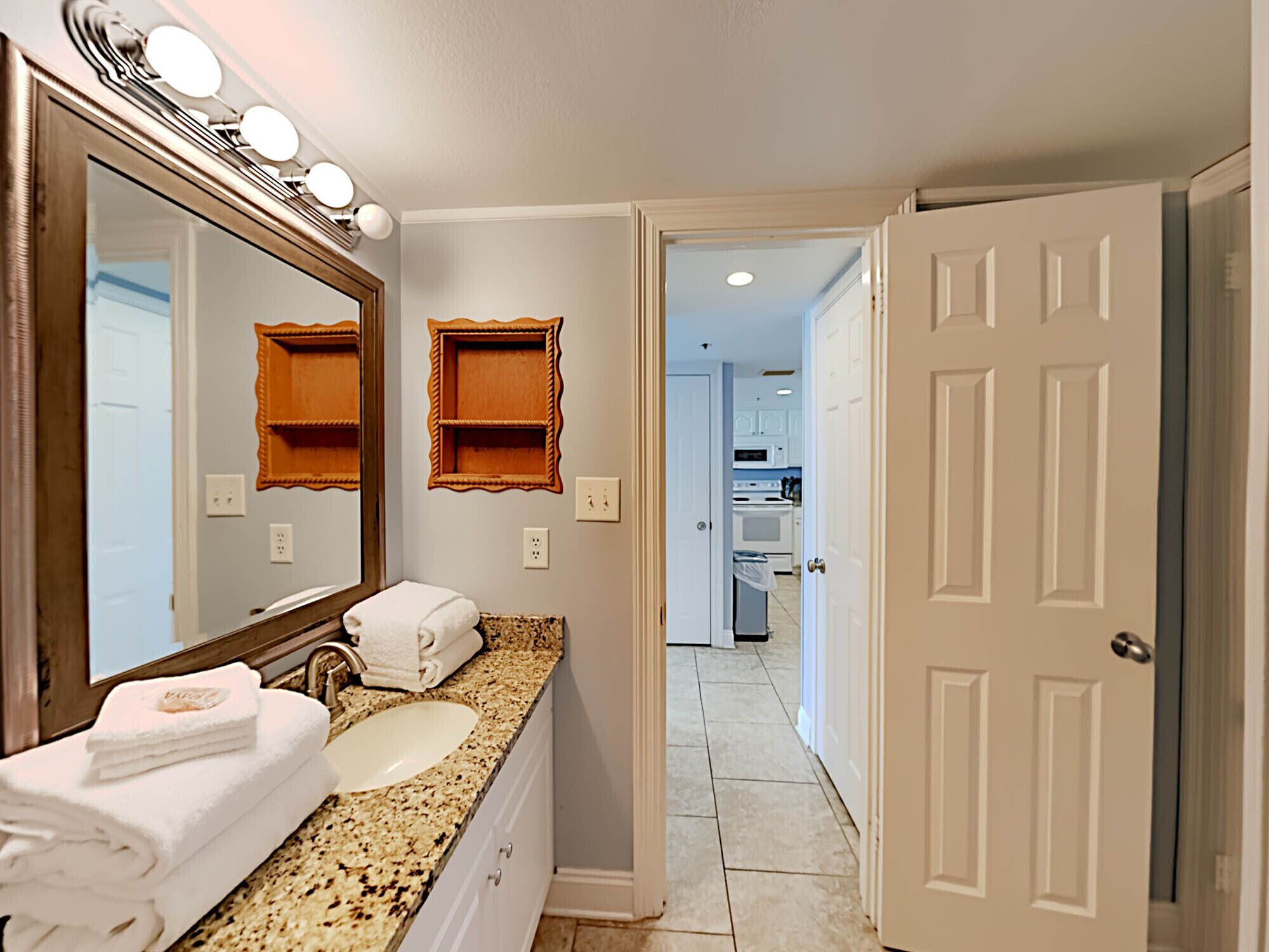 A bathroom with a large mirror, two sinks, and a vanity. There are towels stacked on the counter and a door leading to the hallway.