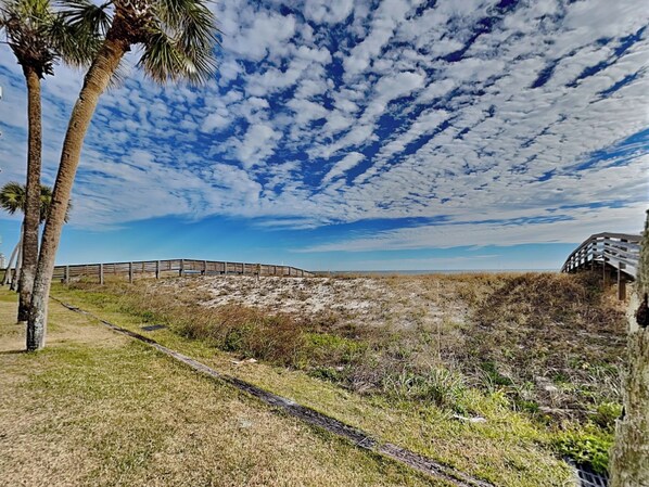 A coastal pathway with a wooden boardwalk, palm trees, and a dune landscape under a partly cloudy sky.