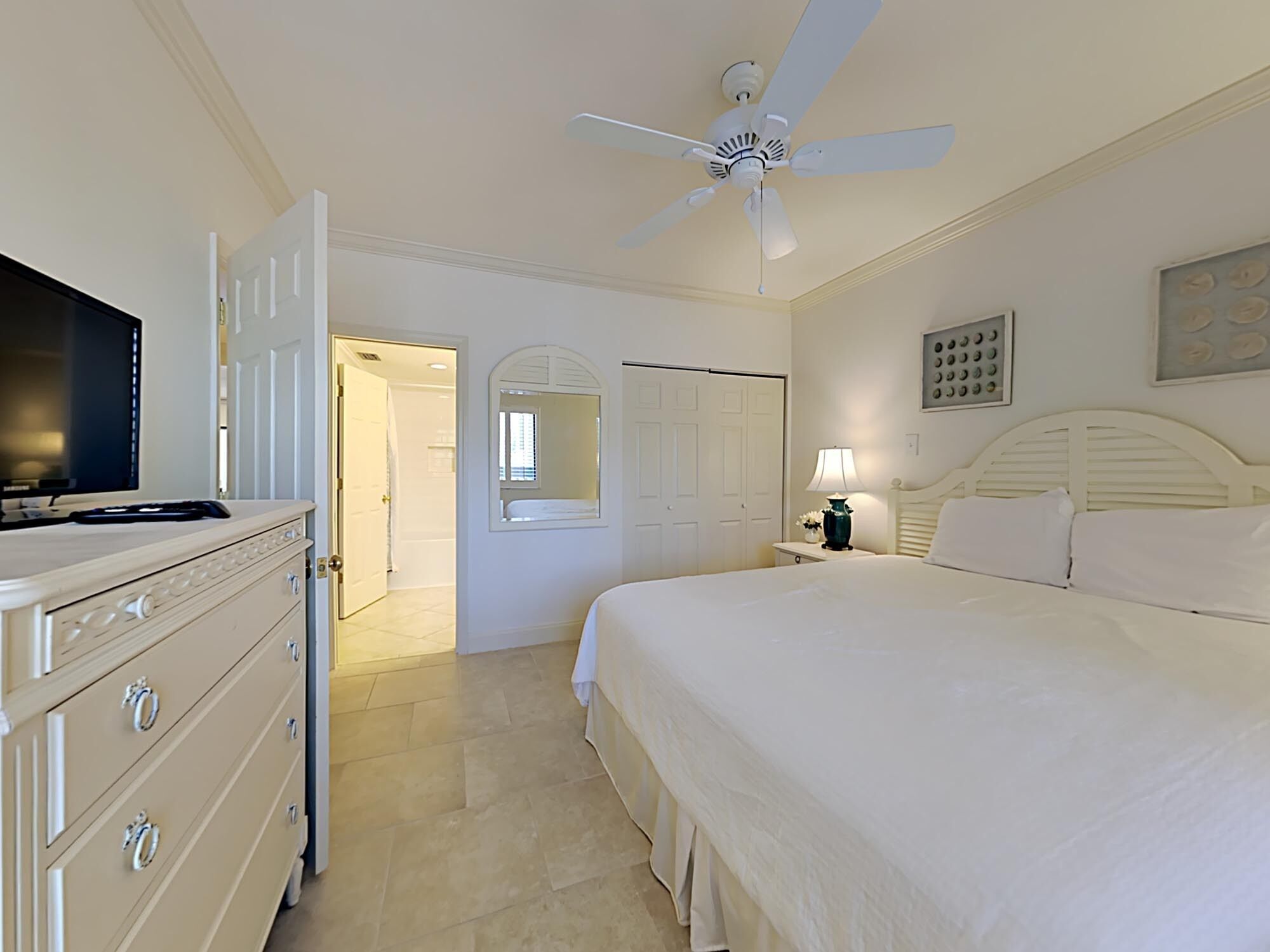 A bedroom with a bed, a dresser, a ceiling fan, a TV, and a door leading to another room.