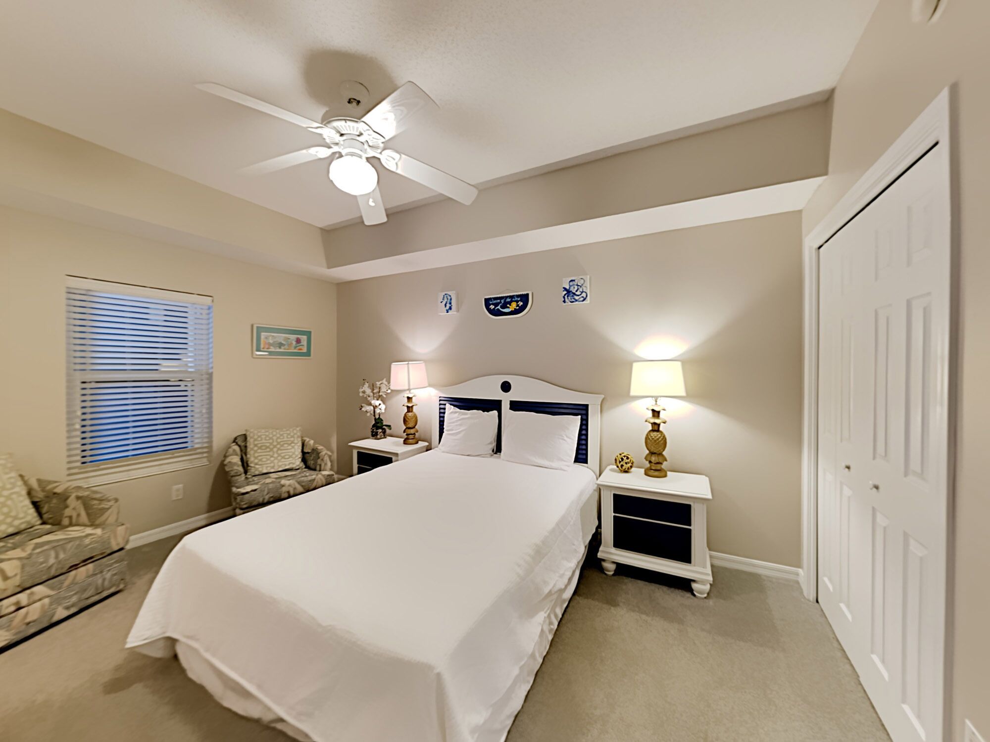 A bedroom with a bed, two bedside tables with lamps, a ceiling fan, a window with blinds, a door, and a wall with decorative plates.