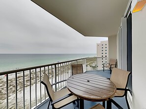 A balcony with a round wooden table and two chairs, overlooking a beach with a clear view of the ocean.