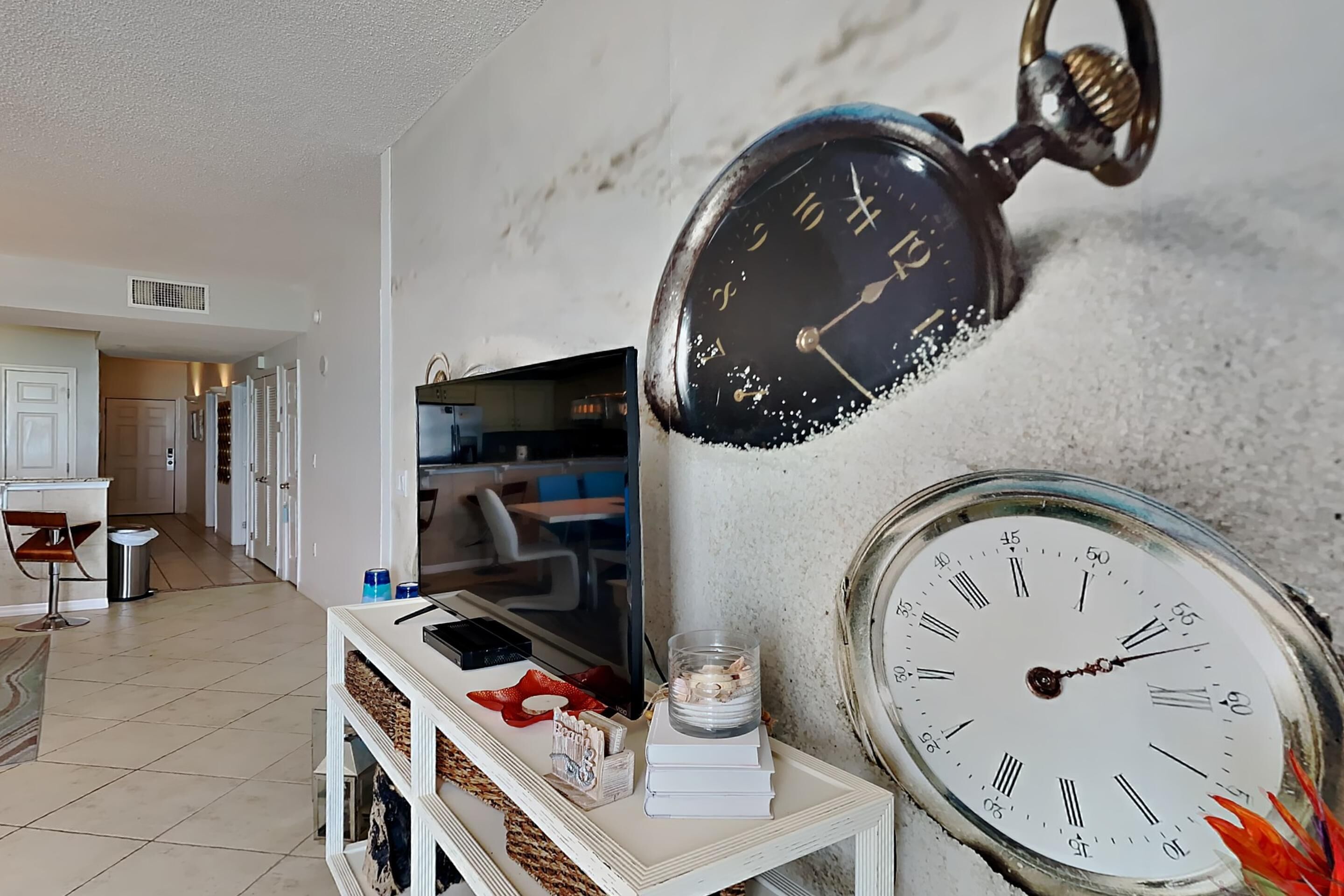Una sala de estar con un televisor sobre un mueble bajo, una consola blanca con objetos decorativos y un reloj de pared.