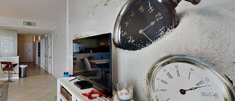 Una sala de estar con un televisor sobre un mueble bajo, una consola blanca con objetos decorativos y un reloj de pared.