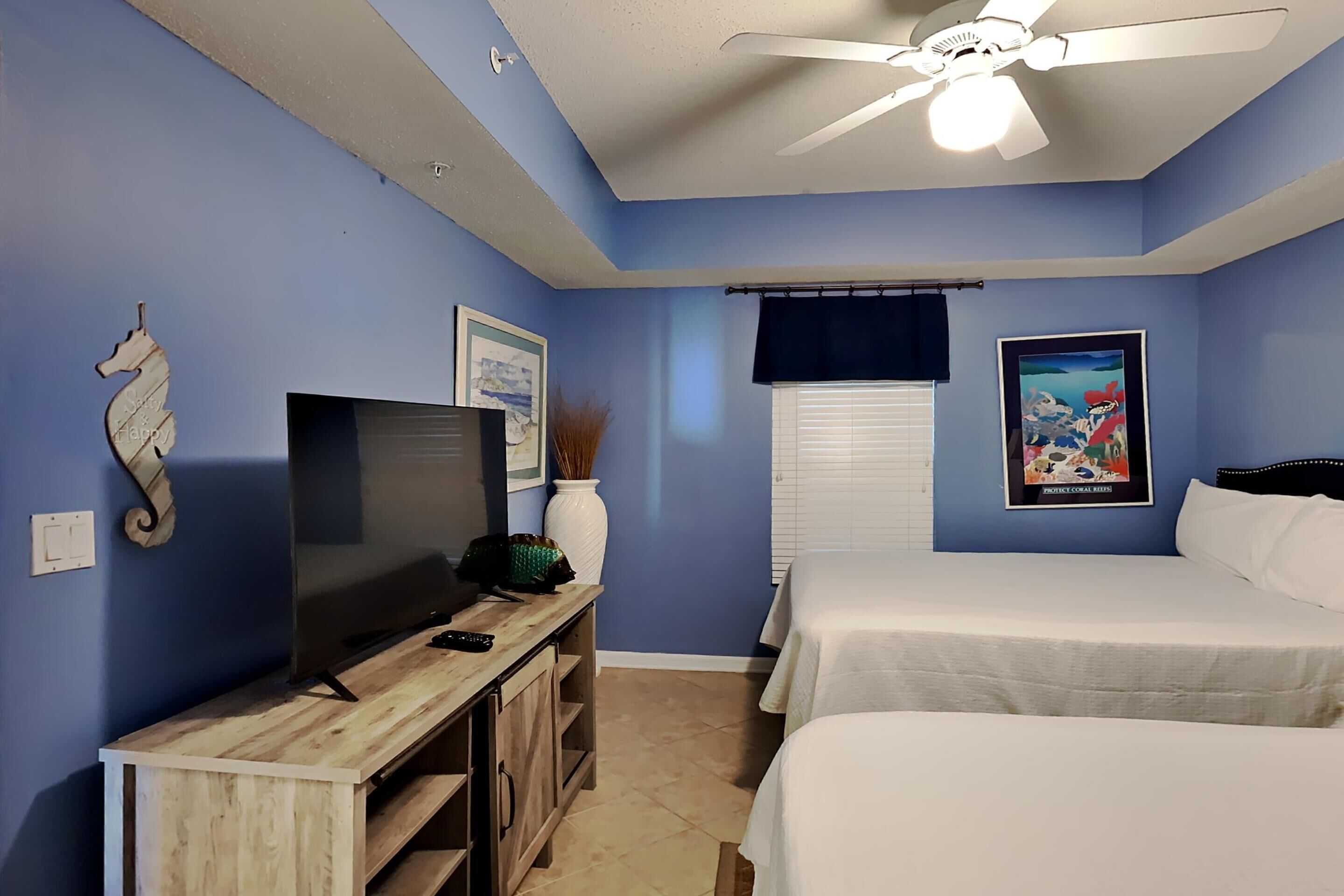 A bedroom with two beds, a TV on a wooden stand, a ceiling fan, and a poster on the wall.