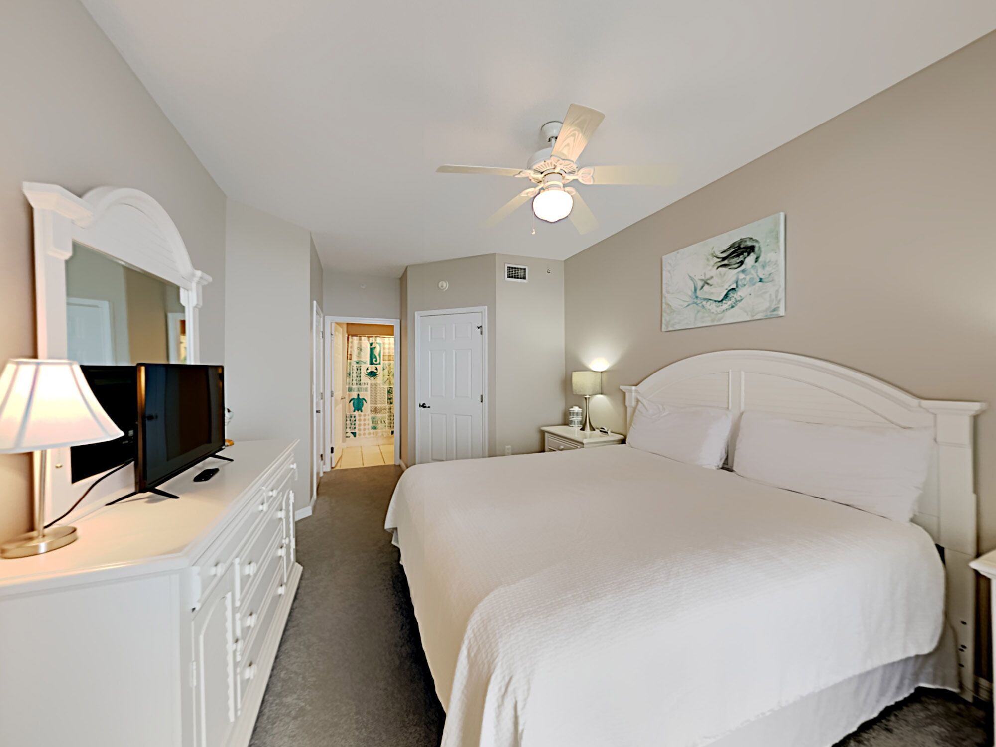 Une chambre d’hôtel équipée d’un lit, d’une télévision, d’un ventilateur de plafond, d’une porte et d’une œuvre d’art accrochée au mur.
