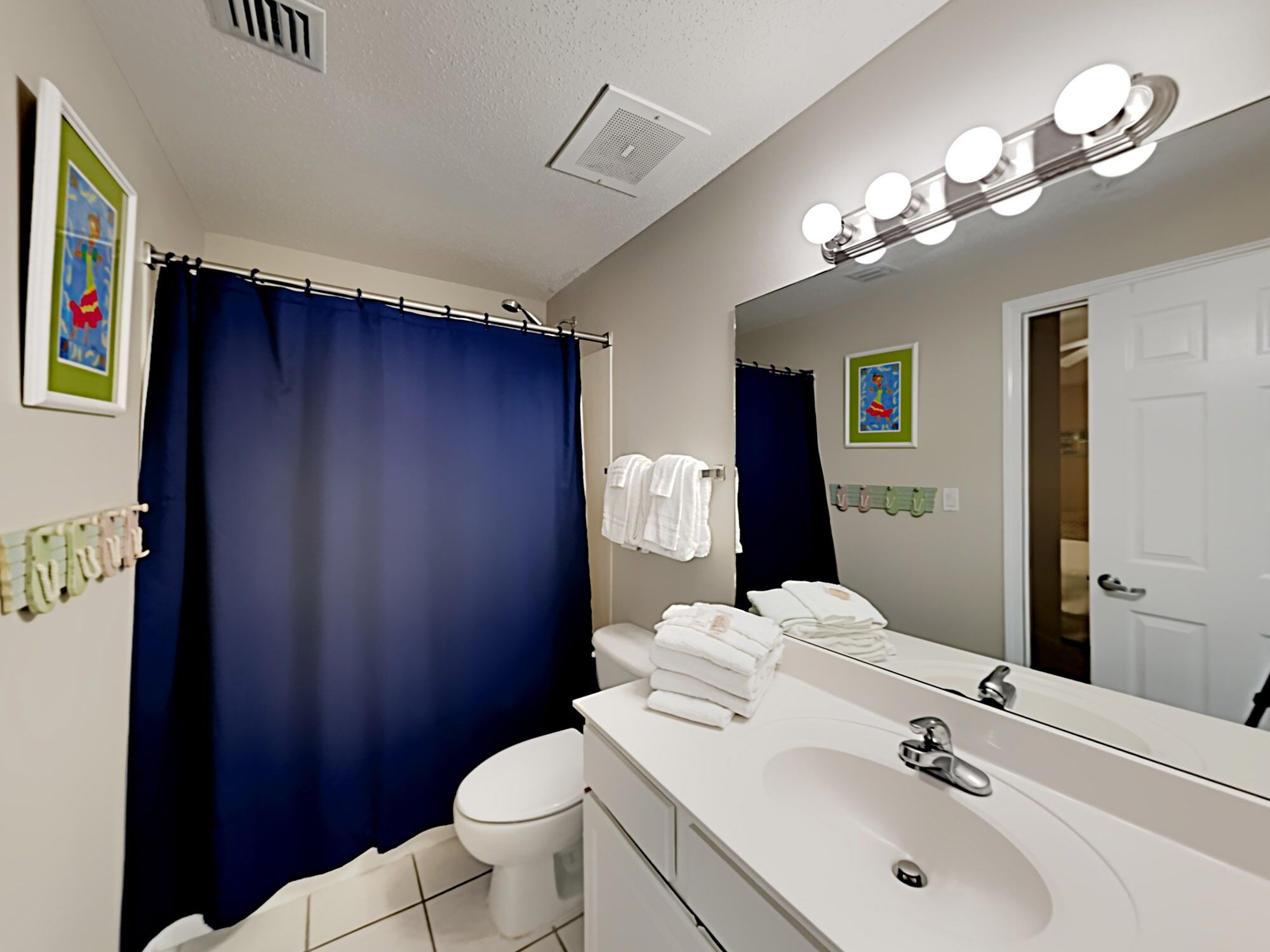 Ein Badezimmer mit einem weißen Waschbecken, einem beleuchteten Spiegel, einem blauen Duschvorhang und einer Toilette.