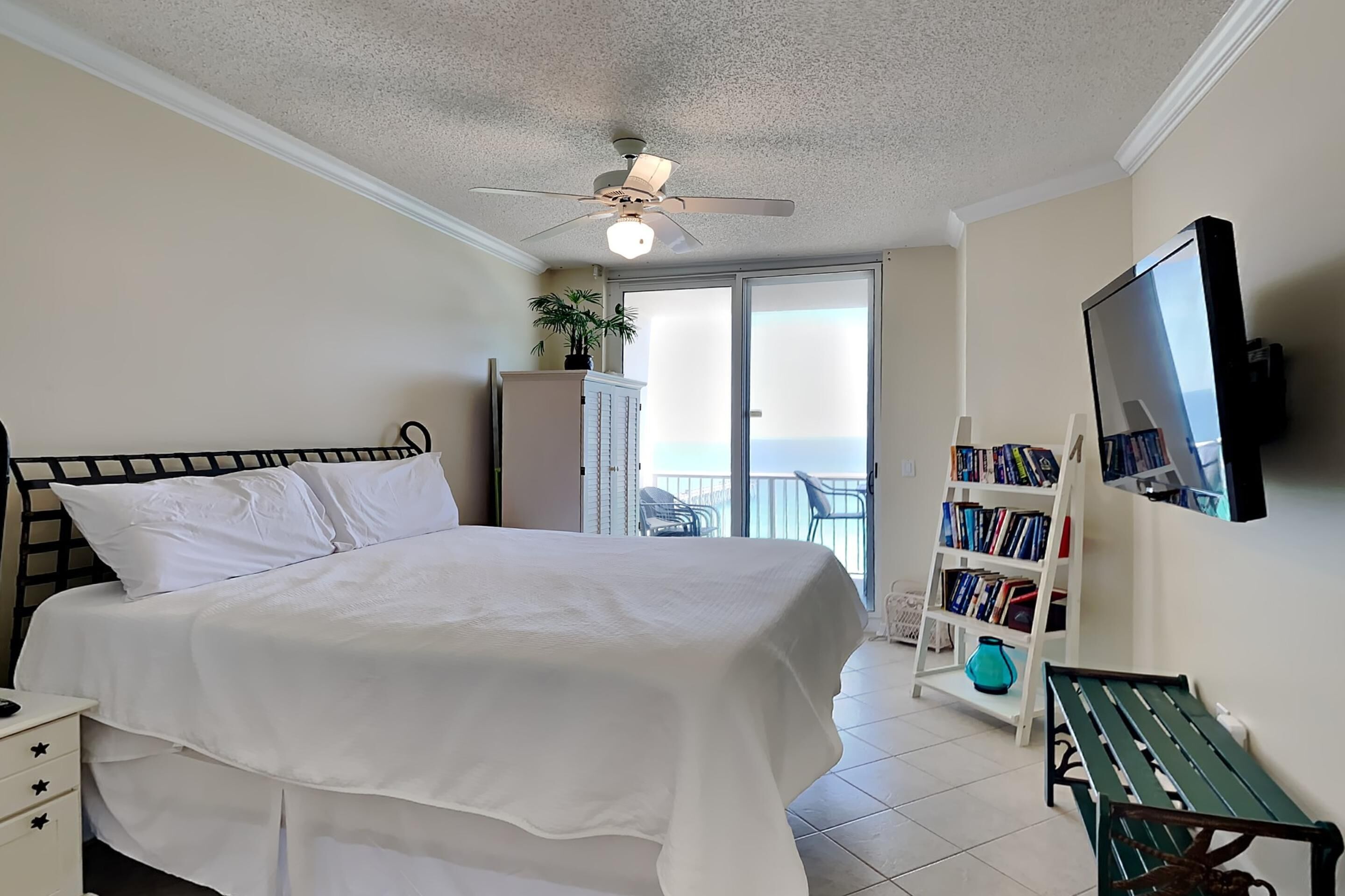 A bedroom with a bed, a ceiling fan, a bookshelf, and a view of the outdoors.