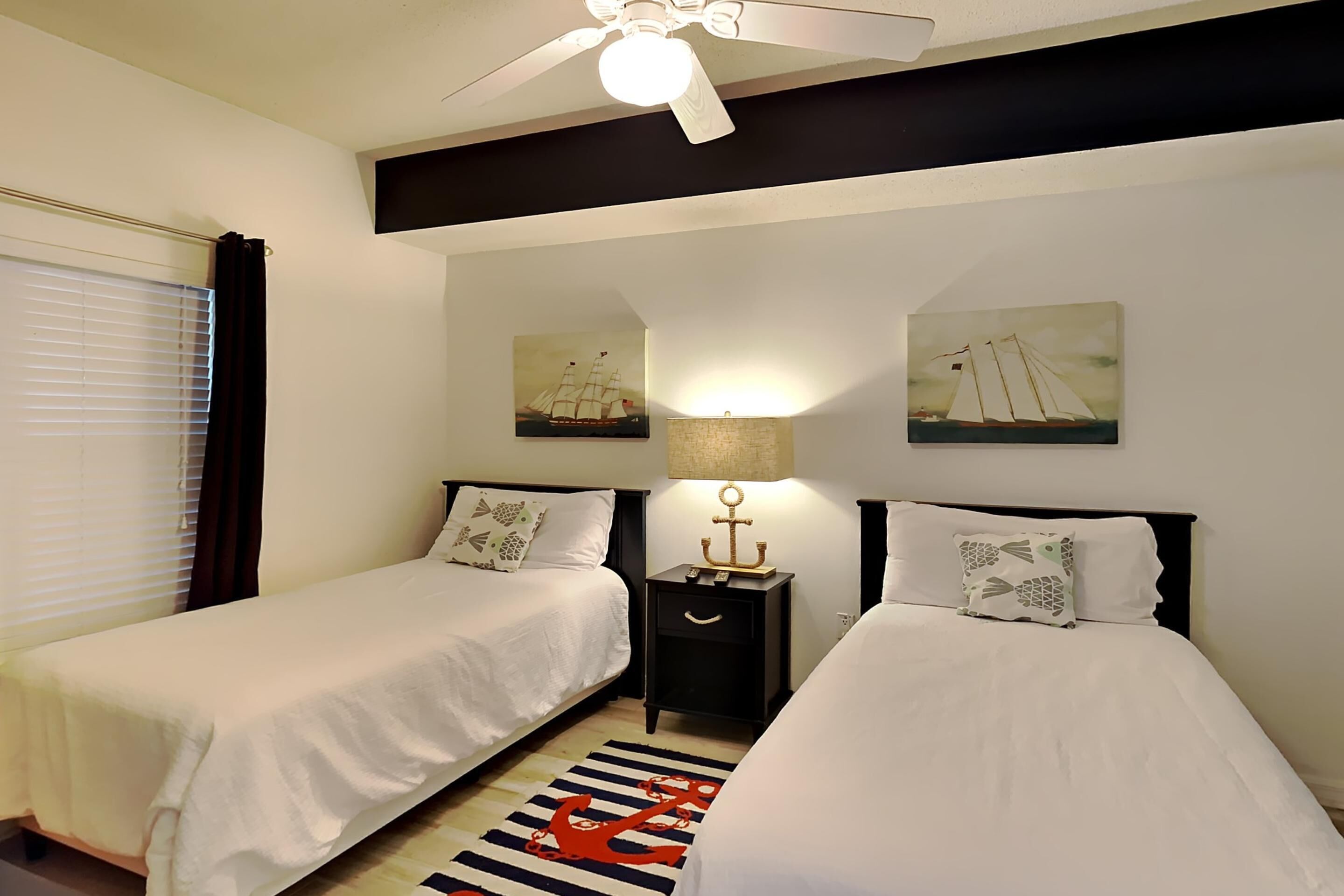 A hotel room with two single beds, a ceiling fan, a nightstand with a lamp, and two wall-mounted paintings of sailboats.