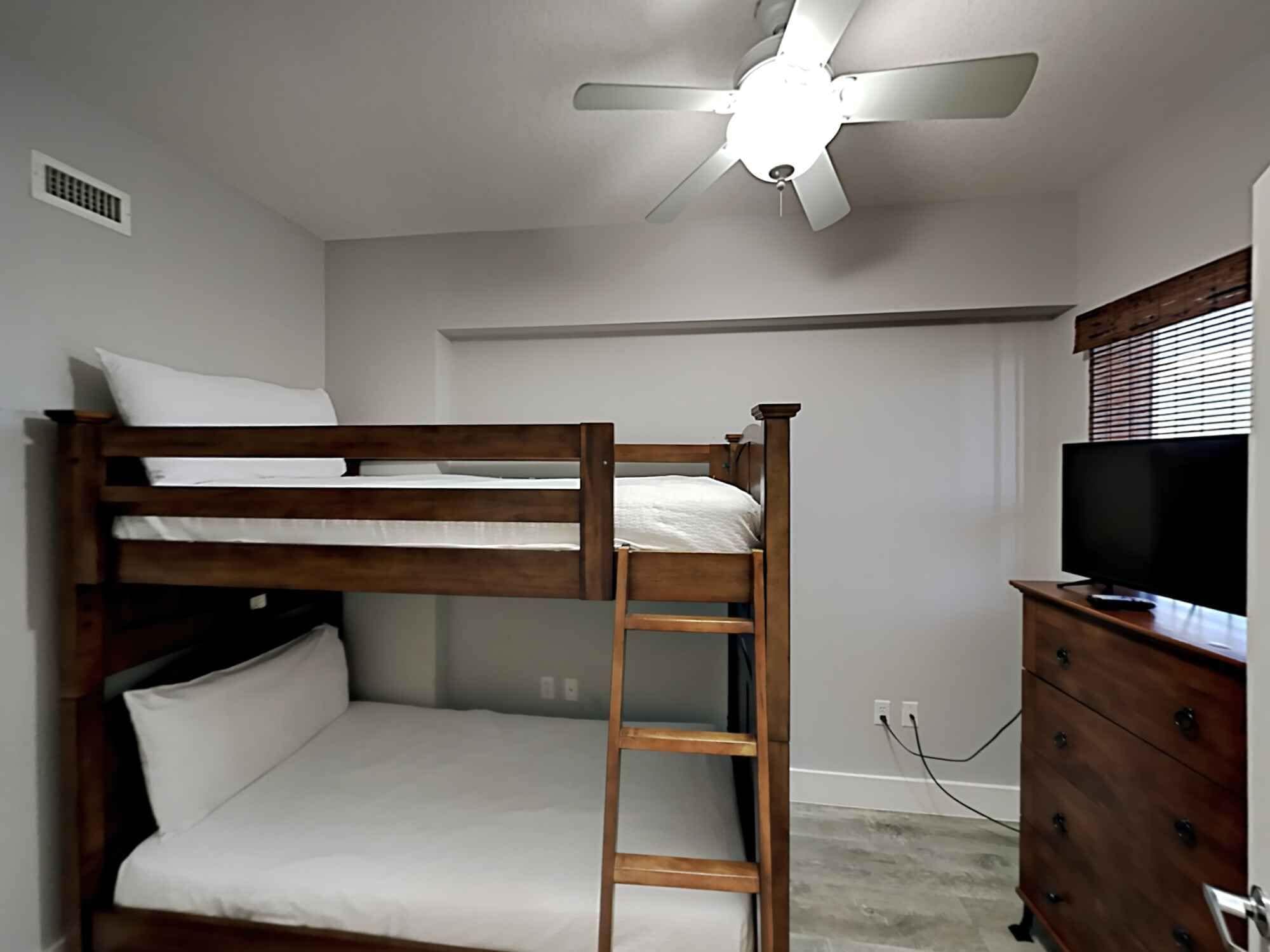 A bunk bed with a ladder, a ceiling fan, a dresser, and a television.