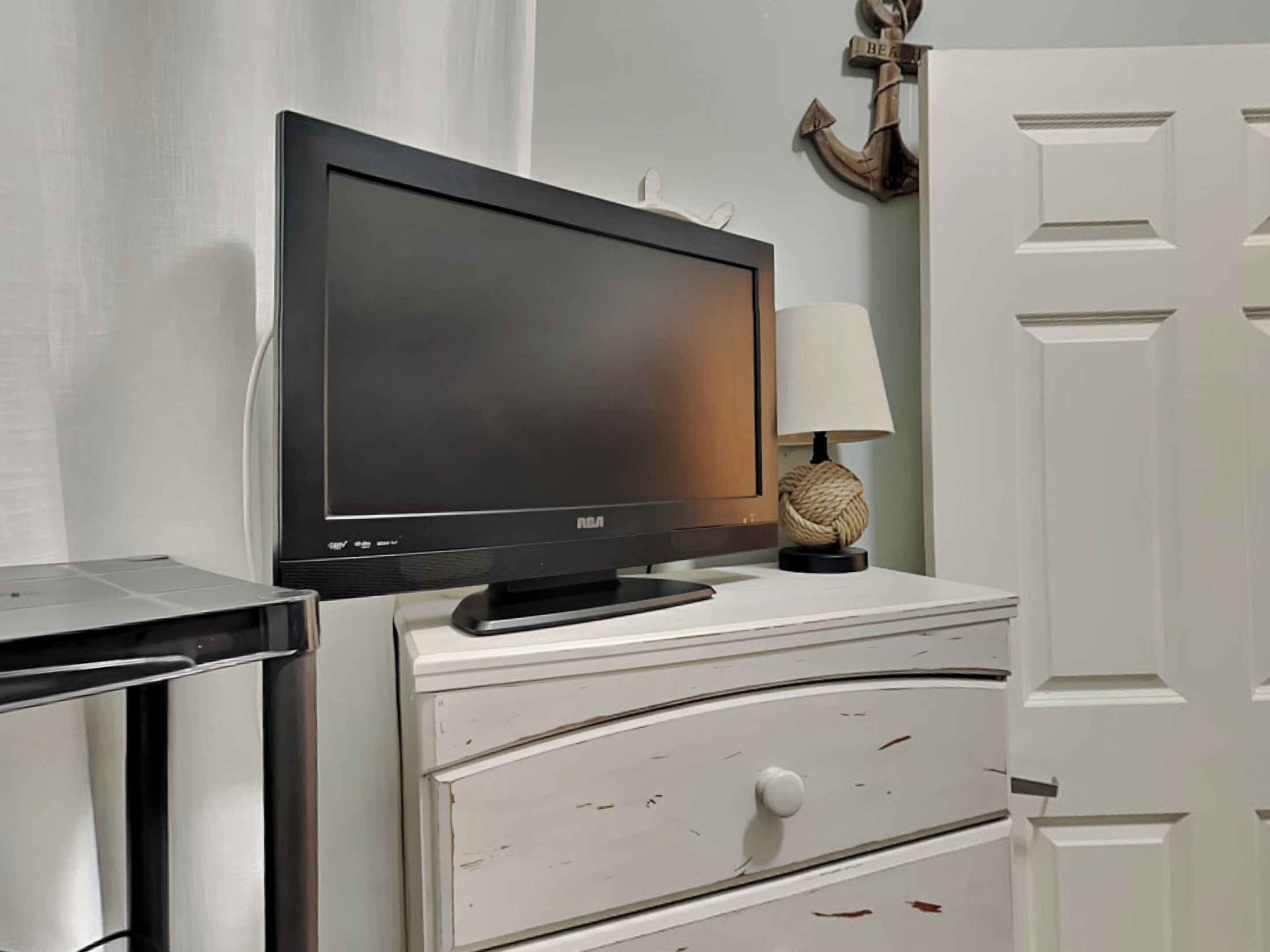 A television on a white dresser, a lamp, and a door with a decorative anchor.