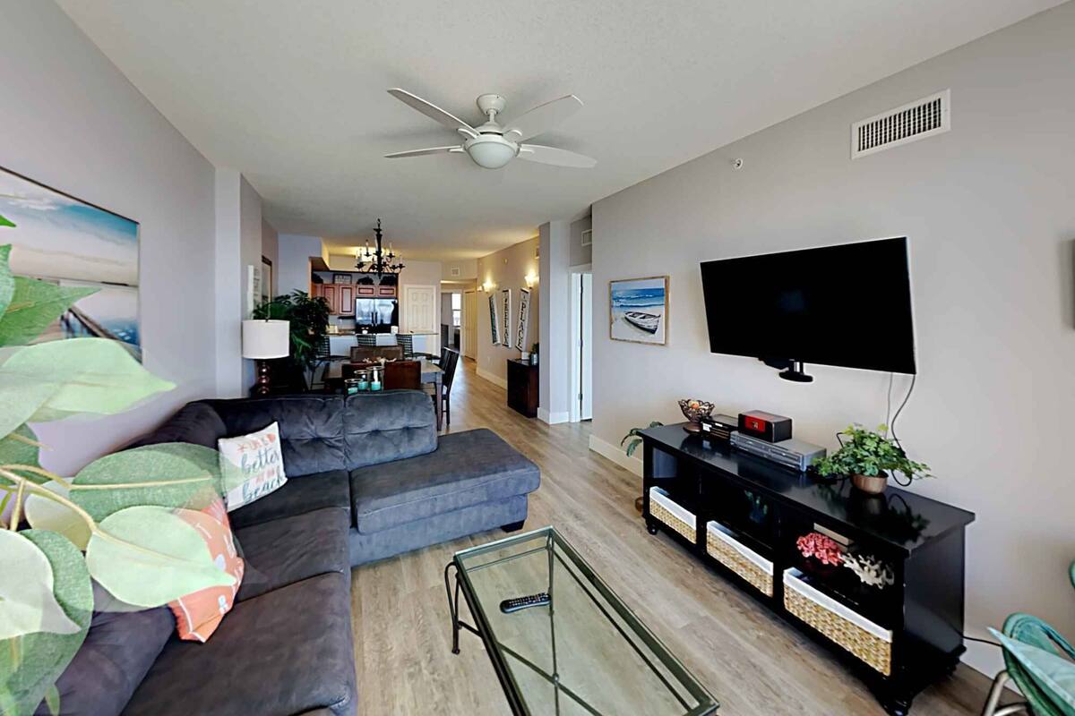 A modern living room with a grey sofa, a glass coffee table, a black entertainment unit, and a flat-screen TV mounted on the wall.